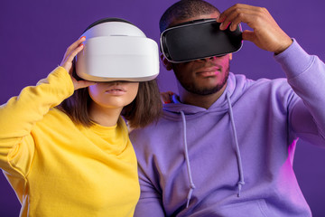 Technology and innovation concept. Two multicultural cheerful people, dressed in yellow and violet coloures, sitting side by side with VR goggles over purple background, having 3d tour