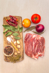 Raw pork shoulder. Spices, dry herbs, onions, garlic, rosemary, bay leaf, nutmeg, pepper mix, tomatoes. Ingredients for marinating meat. Wooden vintage board, stone background, top view, close up.
