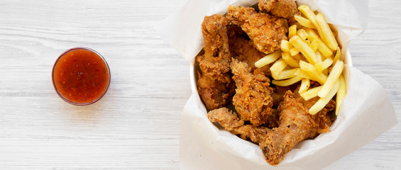 Fried chicken drumsticks, French fries, spicy wings and tenders in paper box and sauce over white wooden surface, overhead view. Flat lay, top view, from above.