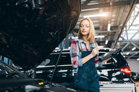 pretty blonde girl working with a computer diagnostic while standing near broken car with opened hood. close up photo. lifestyle
