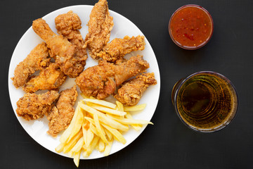 Tasty fastfood: fried chicken drumsticks, spicy wings, French fries, chicken fingers, glass of cold beer, sauce on white plate over black surface, top view. Flat lay, overhead, from above.