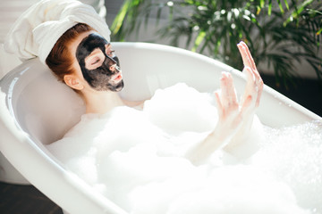 Caucasian young red-haired woman relaxing in bathroom, wearing bath towel wrapped around head, lying in a foam bathtub with black clay face mask on face, enjoying her nail manicure. Body and Skin Care