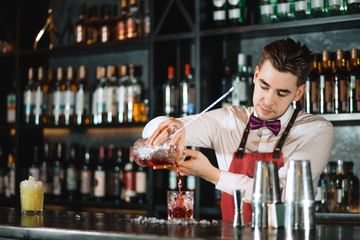 Professional bartender in uniform doing show of his work, holding two parts of metal shaker in his...