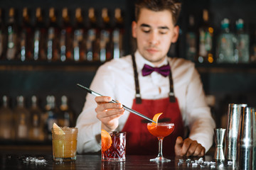 Bartender adding to cocktail in elegant glass one orange zest with tweezers on the bar counter