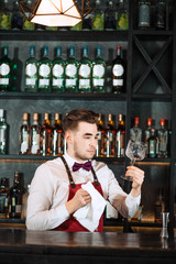 Young handsome smiling barman in bar interior wiping vine glasses. Professional bartender portrait at work in night club cleaning stemware while waiting for orders.