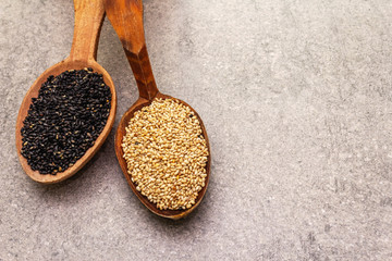 White and black sesame seeds in wooden spoons on stone background, top view, close up.