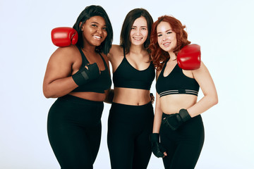 Group of three female boxers posing together in sportswear and red boxing gloves against white background. Multiracial females with different size standing together looking at camera and smiling.