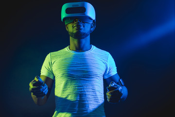 African young man dressed in white t-shirt wearing VR Headset and experiencing virtual reality. Green blue dual color light on dark background.