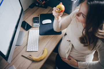 beautiful pregnant brunette woman works at the desk and eats fruits