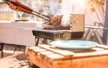 Male chef grill t-bone steak at barbecue dinner outdoor - Man cooking meat for a family bbq meal outside in backyard garden - Summe lifestyle, food and sunday time concept - Focus on hand tongs
