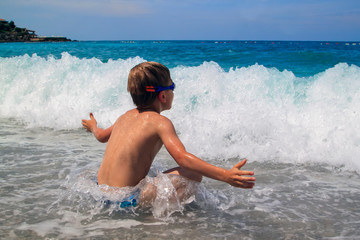 little boy in swimming goggles sitting on pebble on beach and enjoys surging waves with white foam © sommersby