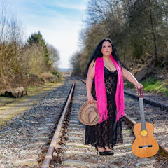 Beautiful mexican woman with black dress standing on the train tracks with a pink shawl, a brown hat and a guitar on an autumn day with blurred background