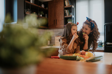 Beautiful woman and her daughter cooking together fresh vegetable salad. Cute girl biting pepper slice, mom biting cucumber.