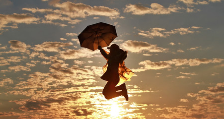 Silhouette of a happy girl with umbrella jumping against the backdrop of an incredible sunset, sky and clouds