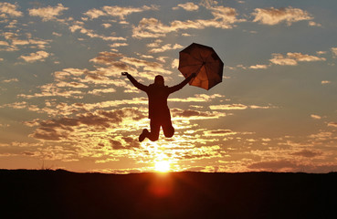 Silhouette of a happy girl with umbrella jumping against the backdrop of an incredible sunset, sky and clouds