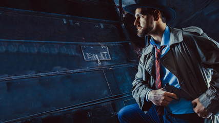 Detective with the gun wearing a fedora hat and a trench coat, dark background.
