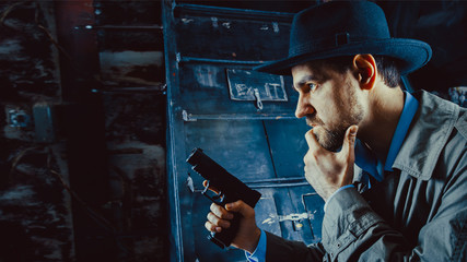 Detective with the gun wearing a fedora hat and a trench coat, dark background. - obrazy, fototapety, plakaty