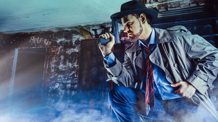 Detective with the gun wearing a fedora hat and a trench coat, dark background.