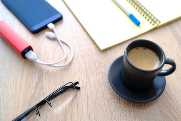 The smartphone is charging from the poverbank. Cup of coffee, diary, pencil and glasses on the table close-up.