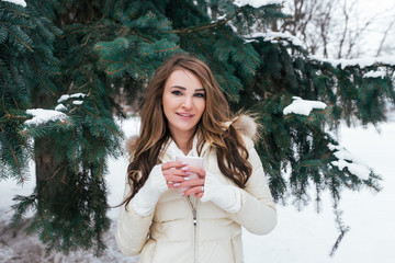 Girl brunette stands in winter in the park on a background of snowy Christmas trees. In his hand holds a mug of hot coffee tea. Warmed in a white jacket happy smiling.