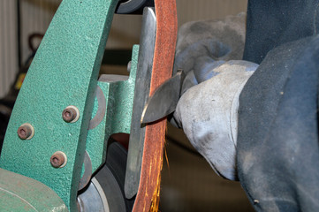 Wearing protective gloves, the professional knife maker keeps safety in mind when using the grinder to sharpen the blade as sparks fly. Bokeh effect.