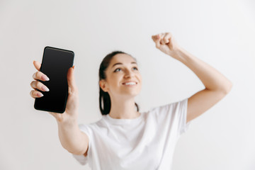 Portrait of a confident casual girl showing blank screen of mobile phone isolated over gray background at studio.