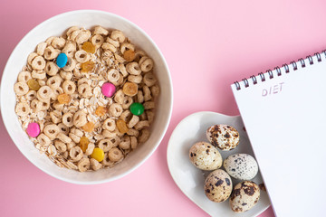 Corn flakes and oatmeal in the white bowl, quail eggs and notepad on pink background . Diet concept.