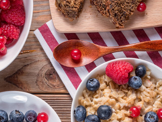 Close up view of wooden spoon between porridge with blueberries and raspberries and grain bread on wooden background. Vegan breakfast.