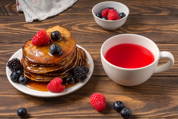 Pancakes covered by honey with blueberries and strawberries and cup of red juice on wooden background