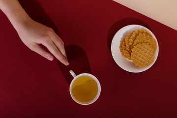 Obraz na płótnie Canvas Ceramic cup with tea and waffle, on a red background. The harsh shade of the sun, the art concept of the morning and the charge of energy. Girl hand reaches for the mug.