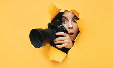 A paparazzi funny girl with a camera looks out from cover and looks at what is happening with her mouth open. Yellow paper, torn hole. Tabloid press. In search of the plot for photo stocks. - obrazy, fototapety, plakaty