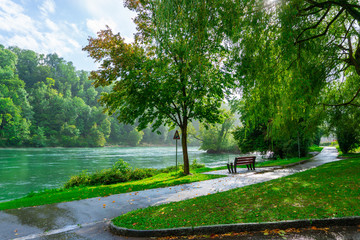 Fototapeta na wymiar Landscape of a park with wooden bench and green leave trees and river with reflection of green trees in rainy day