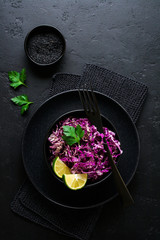 Salad of purple Peking cabbage with olive oil, lime and black sesame seeds in ceramic plate on dark concrete background.