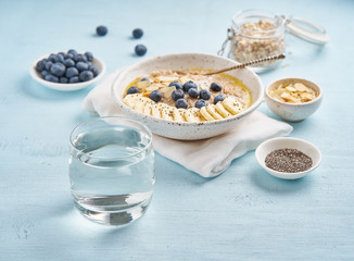 Glass of clean water and healthy diet breakfast with oatmeal, blueberries, banana on blue light background. Side view.