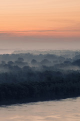 Mystical view on riverbank of large island with forest under haze at early morning. Eerie mist among layers from tree silhouettes. Morning atmospheric landscape of majestic nature in blue faded tones.