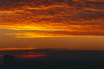 Obraz premium Cityscape with vivid fiery dawn. Amazing warm dramatic cloudy sky above dark silhouettes of city building roofs. Orange sunlight. Atmospheric background of sunrise in overcast weather. Copy space.