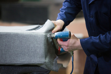 Closeup of a young man in a furniture factory who puts together one part of the sofa with a stapler - obrazy, fototapety, plakaty