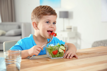 Adorable little boy eating vegetable salad at table in room