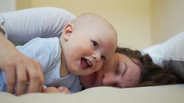 Cheerful baby at the age of one year old is lying in bed with her mother and laughing