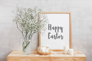Frame with text HAPPY EASTER, glass vase with gypsophila flowers, nest with eggs, feathers and candles on a wooden table on a background of light gray walls. Easter home decor. Style room interior.