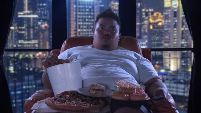 Unhealthy lifestyle concept. Greedy overweight man watching TV while eating junk foods on the sofa in the apartment at night. Shot in 4k resolution
