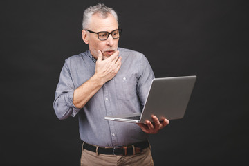 OMG! I'm shocked! Scared senior aged bearded old man in eyeglasses holding laptop computer isolated against black background.