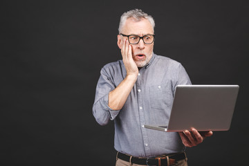 OMG! I'm shocked! Scared senior aged bearded old man in eyeglasses holding laptop computer isolated against black background.