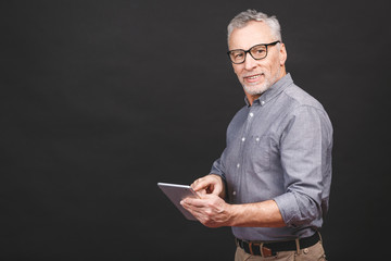 Great news! Handsome mature businessman in smart casual wear and eyeglasses is using a digital tablet and smiling, isolated against black background. Looking at camera.