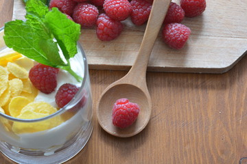 a breakfast glass of muesli and greek yoghurt with raspberries next to a chopping board with raspberries on top on a wooden table