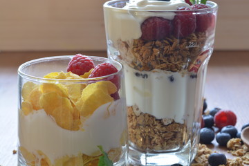 a breakfast glass with granola and and a breakfast glass with muesli and greek yoghurt in front of a wooden background