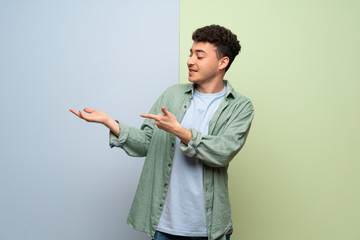 Young man over blue and green background holding copyspace imaginary on the palm to insert an ad
