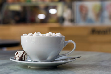 Mug of coffee with marshmallows on porcelain plate on blurred colorful interior bokeh background.