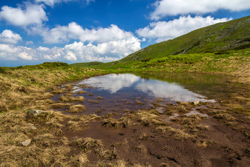 Obraz premium Blue sky with bright white clouds reflected in small lake between green hills on sunny day. Summer panorama.