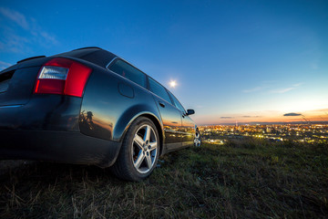 Plakat Black car parked at night in green meadows on copy space background of lights of distant city buildings and bright blue sky with first star at sunset.